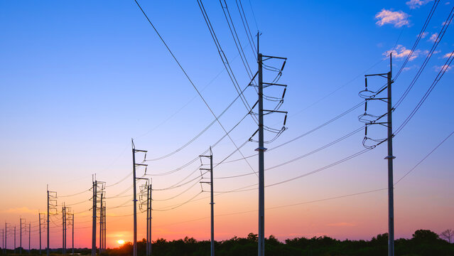 Silhouette electric poles with cable lines on country road with golden sun on blue sunset sky background, low angle and perspective side view