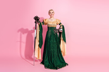 Fototapeta premium Elegant young woman in medieval attire with knightly gauntlet and sword posing against a pink background