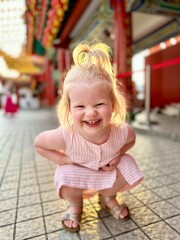 Petite fille blonde vêtue de rose souriant accroupie sur fond de temple asiatique en Malaisie