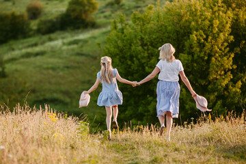 Fototapeta premium Middle-aged woman and young girl in blue dresses holding hands and walking through a scenic green field.