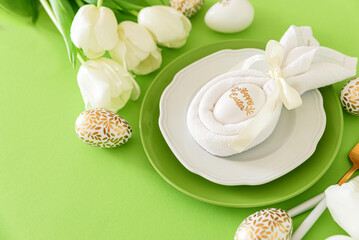 Plate decorated bunny ear napkin, white tulips and Easter eggs on green background. Easter celebration concept. Flat lay. Top view