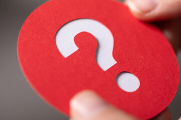 Close-up of a hand holding a red circle with a white question mark cutout. Uncertainty concept.