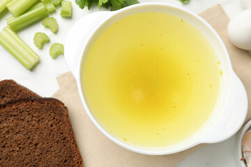 Tasty chicken bouillon in bowl with bread and ingredients on white table, flat lay