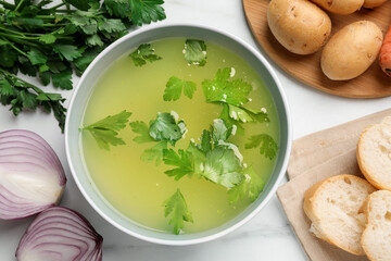 Tasty chicken bouillon in bowl, bread and ingredients on white marble table, flat lay