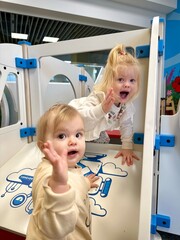 Deux sœurs blonde jouant sur une aire de jeux intérieure faisant signe bonjour © Mathieu