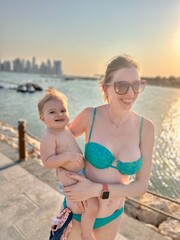 Jeune femme en maillot de bain turquoise de type bikini portant un bébé devant la mer sur fond de...