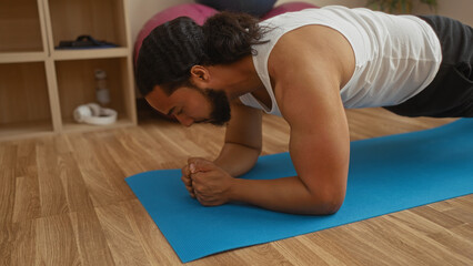 Man exercising indoors on a yoga mat at a wellness center, displaying focus and strength in a plank position, surrounded by spa-like calming environment.