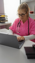 Woman working on laptop in clinic wearing pink scrubs and stethoscope, focusing intently indoors at...