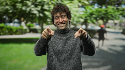 Young hispanic man pointing at camera outdoors in a park setting, exuding confidence and charisma against a lush green background.