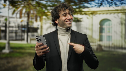 Young hispanic man points at smartphone while smiling outdoors on a city street showing technology...