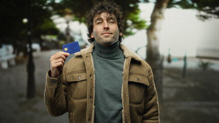 Fototapeta premium Young hispanic man outdoors holding credit card in a city street wearing brown jacket and looking confident with blurred background enhancing the urban setting.