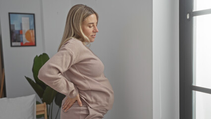 Pregnant woman with blonde hair stands indoors, resting hands on her back, in a cozy bedroom setting, highlighting her expecting belly and creating a serene home atmosphere.