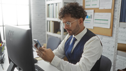 Fototapeta premium Young man focuses intently on smartphone screen while working at desk in modern office, surrounded by computer and bookshelves.