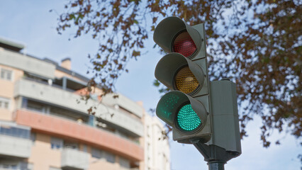 Traffic light with glowing green signal against the backdrop of a city building and tree foliage in...