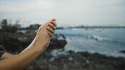 Man clapping hands with fingers intertwined focused near seaside with ocean waves crashing on rocky shore under clear blue sky during bright daylight hours
