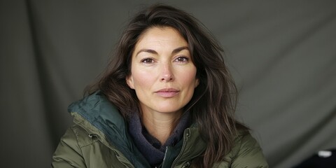 Fototapeta premium A woman with long, loose hair sits comfortably, wearing a warm green jacket. She looks directly at the camera with a striking expression, suggesting warmth and confidence