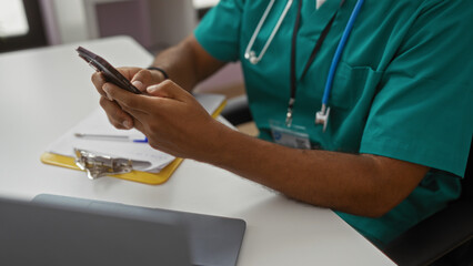 Man texting in scrubs in a clinic room with a clipboard, pen, stethoscope, and badge, highlighting a professional medical setting indoors.