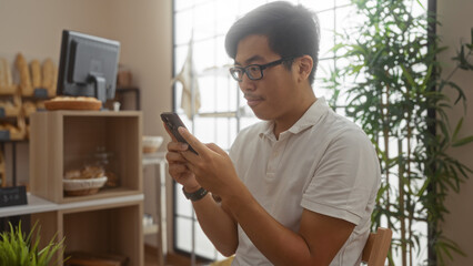 Fototapeta premium Young man in a modern bakery cafe using a smartphone with a focused expression against a background of bread and greenery