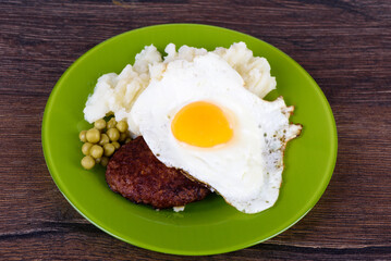 Freshly cooked cutlet with fried egg and mashed potatoes with green peas on wooden background.