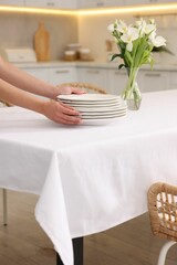 Woman putting plates on table with white tablecloth in kitchen, closeup