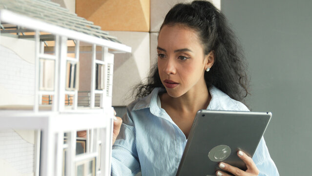 Young beautiful hispanic architect engineer drawing building construction by using tablet while checking house model structure at modern office. Creative business construction concept. Manipulator.