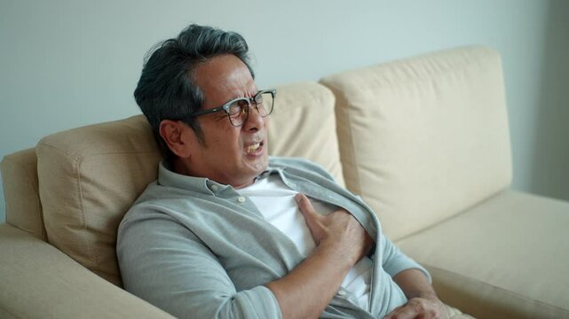 Asian senior mature man suffering chest pain from heart attack while sitting alone on sofa in living room at home, old male having cardiac disease problem, medical insurance for retirement concept.
