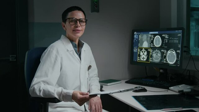 Medium portrait of cheerful, confident Caucasian female radiologist in white coat, glasses, at workplace in clinic, sitting at desk, in front of computer with patient brain scan, looking at camera