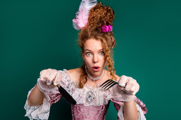 Elegant young woman in antique pink dress holding cutlery with surprise in a vibrant green...