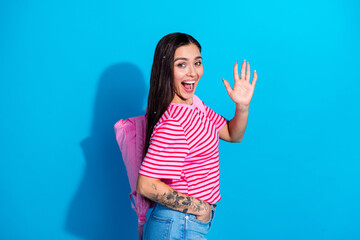 Fototapeta premium Smiling Young Woman with Pink Backpack Waving on Blue Background Wears Striped Shirt