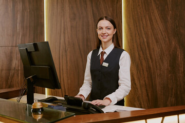 Medium close up of adorable smiling receptionist standing near table with computer while working at...