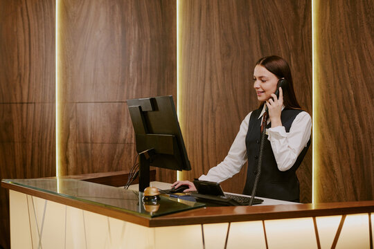 Wide shot of young adult woman looking at computers screen and answering phone call while working as receptionist at hotel