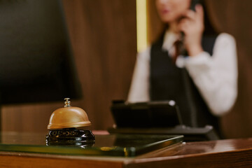 Medium close up of golden bell on polished table in hotel lobby, unrecognizable receptionist peaking on phone in blurred background