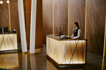 Wide shot of luxurious hotel lobby with adorable young adult Caucasian receptionist working on...
