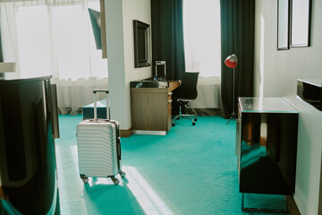 Empty spacious hotel room with turquoise carpet, wooden furniture and silver suitcase