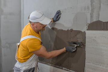 Tiling with large format tiles. A tiler uses a notched trowel to spread tile adhesive onto a wall.