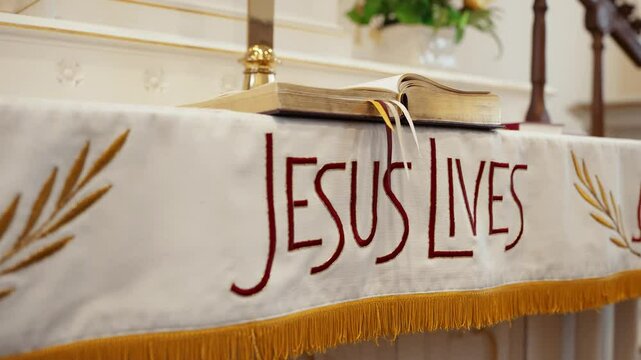 A white cloth that says Jesus Lives under The Holy Bible in a small church chapel.