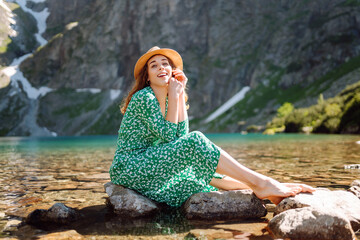 Young woman enjoying view of mountain lake in sunbeams. Beautiful woman in stylish dress traveling in mountains. Adventure concept, tourist, vacation.