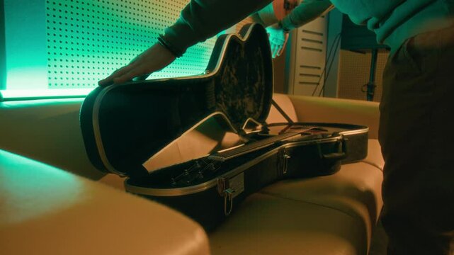 Side midsection shot of impersonal musician putting acoustic guitar in black case on sofa, closing locks and taking it away in neon lit room