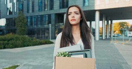 Woman walks in front of glass modern office building of corporation where she worked being laid off from her position girl with sad uncertain look on face holding box of packed belongings unemployment