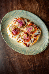 A delicious dish featuring a pizza topped with melted cheese, pickles, tomatoes, red onions, and a savory sauce, presented on a light green patterned plate against a wooden background.
