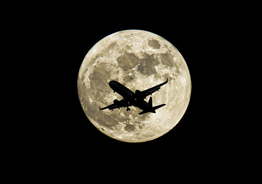 Silhouette of a plane flying in front of a large moon and dark sky background.