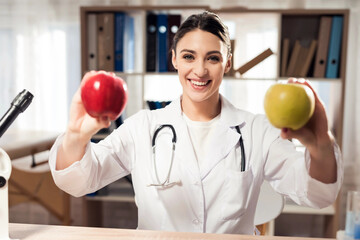 Portrait of smiling female doctor with stethoscope holding red and yellow apples isolated.