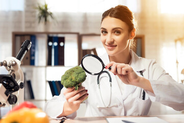 Female doctor sitting at desk in office with microscope and stethoscope. Woman is holding broccoli...