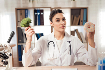 Female doctor sitting at desk in office with microscope and stethoscope. Woman is holding broccoli...