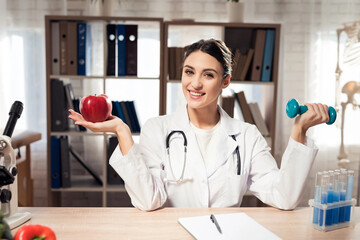 Female doctor sitting at desk in office with microscope and stethoscope. Woman is holding apple and...