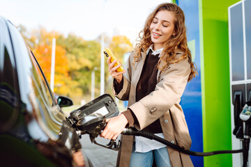 A young woman at a gas station fills up her car with one hand and checks her smartphone with the...