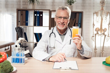 Doctor sitting at desk in office with microscope and stethoscope. Man is holding juice.