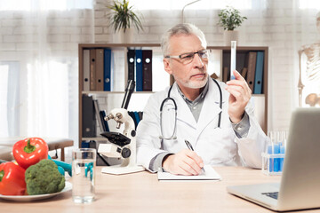 Doctor sitting at desk in office with microscope and stethoscope. Man is holding beaker.