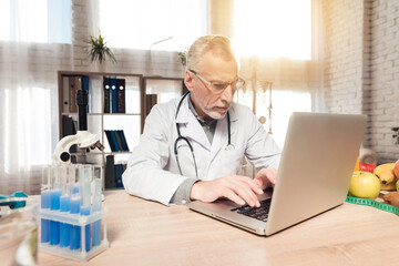 Doctor sitting at desk in office with microscope and stethoscope. Man is working on laptop.
