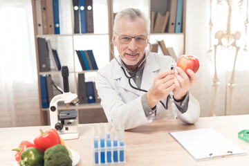 Doctor sitting at desk in office, listening apple with stethoscope.